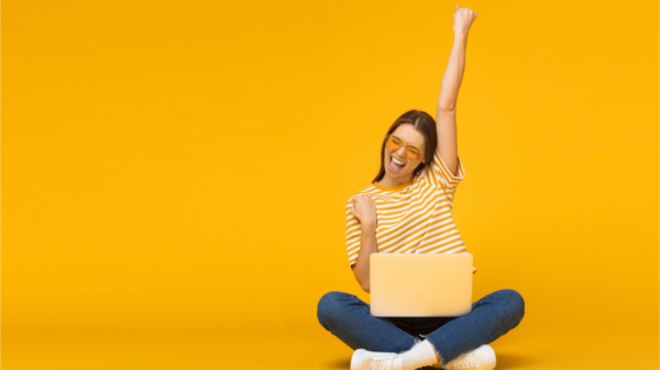 Garota sorridente animada sentada no chão com o laptop, levantando uma mão no ar, isolada no fundo amarelo.