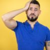 Homem com barba, vestindo camisa polo azul sobre fundo amarelo, colocando uma mão na cabeça e sorrindo como se tivesse esquecido algo.