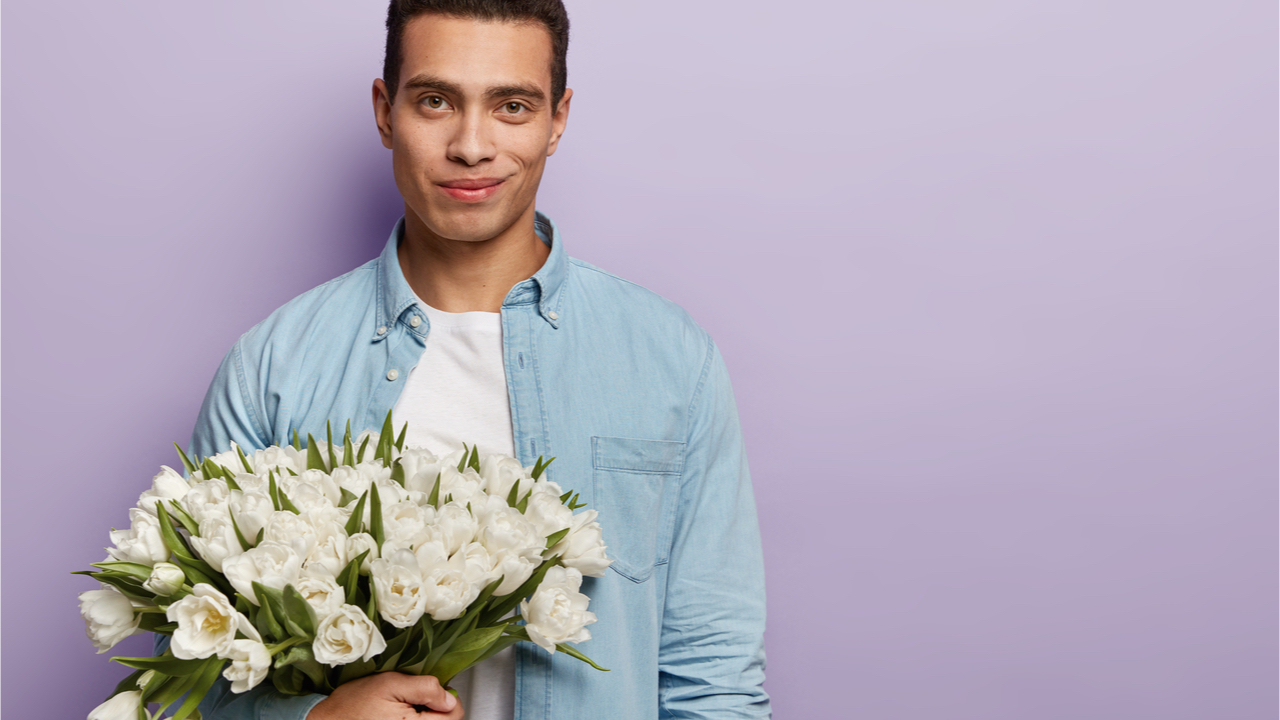 Rapaz com buquê de flores brancas esperando namorada, indo fazer a proposta e confessar o amor, com um olhar atraente e um sorriso gentil, isolado sobre a parede roxa do estúdio.