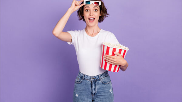 Retrato fotográfico de menina sorrindo, segurando caixa de pipoca, tirando óculos 3d, assistindo filme, isolada em fundo de cor roxa vibrante.