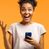 Garota espantada segurando smartphone, feliz em receber notificações, gesticulando felicidade ativamente. Foto de garota em fundo laranja. Conceito de emoções e sentimentos agradáveis.