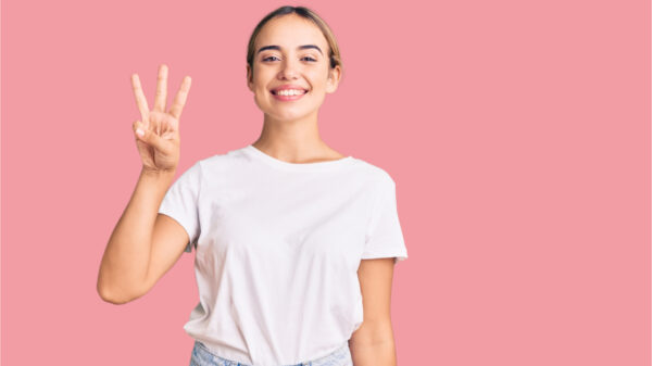 Jovem mulher loira vestindo camiseta branca casual, mostrando e apontando para cima com os dedos em número três enquanto sorri confiante e feliz.