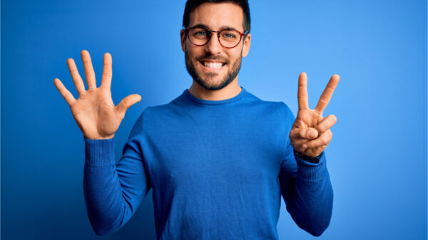 Jovem com barba, vestindo suéter casual e usando óculos sobre fundo azul, mostrando e apontando para cima com os dedos fazendo o número sete enquanto sorri confiante e feliz.