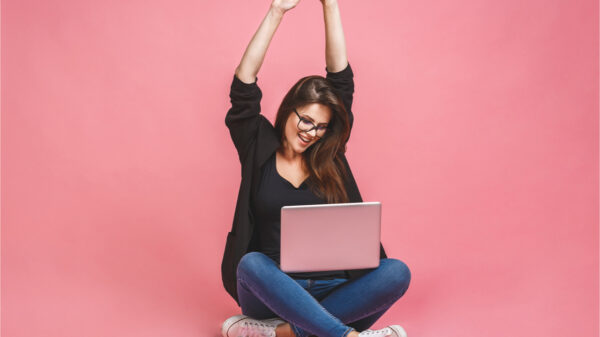 Retrato de mulher feliz, sentada no chão em pose de lótus e segurando laptop, isolada sobre fundo rosa.