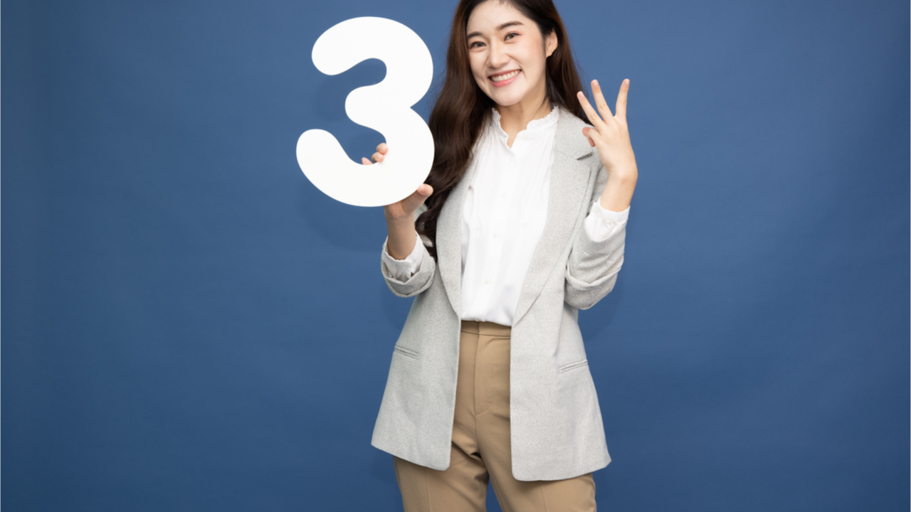 Jovem mulher mostrando o número 3 e apontando para cima com o dedo em número 3, isolada em fundo azul.