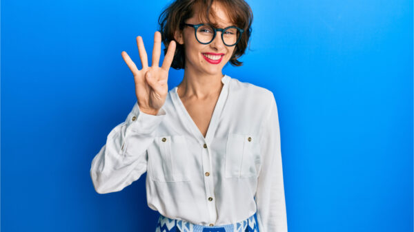 Jovem morena vestindo roupas casuais e óculos, mostrando e apontando para cima com os dedos em número 4 enquanto sorri confiante e feliz.