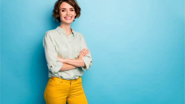 Foto de mulher de negócios com penteado curto, amigável, radiante, sorridente, de braços cruzados, de bom humor, vestindo camisa verde casual e calça amarela, isolada no fundo de cor azul.