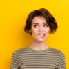 Foto aproximada de uma garota insegura, olhando para o lado, mordendo os lábios, isolada em fundo amarelo brilhante.