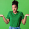 Retrato de uma jovem encolhendo os ombros, olhando para a câmera posando sobre fundo verde. Foto de estúdio. Mulher intrigada.