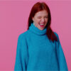 Adolescente ruiva sorrindo e feliz, de suéter azul, piscando o olho, olhando para a câmera dando um sorriso, piscando e flertando, expressando otimismo. Jovem mulher adulta no fundo do estúdio rosa.