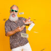 Retrato de homem engraçado, velho, barbudo e milionário de óculos desperdiçando dinheiro, jogarndo notas, usando shorts e camisa de leopardo, isolado sobre fundo amarelo.