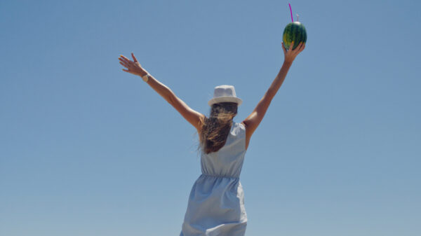 mulher na praia com as mãos ao alto segurando uma água de coco