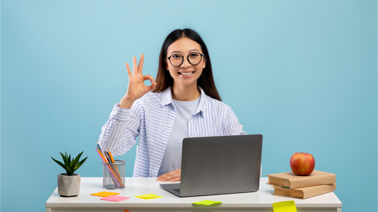 Mulher feliz, de óculos, mostrando gesto de sinal bem, sentada na mesa e usando o laptop sobre fundo azul.