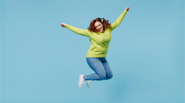 Mulher jovem, alegre e animada vestindo blusa verde, dando um salto alto com as mãos estendidas, isolada no no fundo azul liso. Conceito de estilo de vida de pessoas.