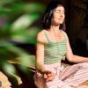 mulher usando blusa verde e fazendo meditação em um ambiente iluminado em com plantas