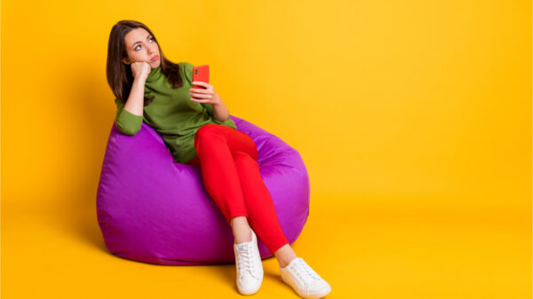 Foto de uma mulher triste e entediada, sentada no puff, segurando celular, isolada no fundo de cor amarela.