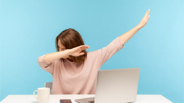 Mulher muito feliz mostrando gesto de dança "dab", realizando meme de sucesso na internet, sentada no local de trabalho com laptop, em escritório em casa. Foto de estúdio interno isolado em fundo azul.