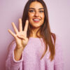 Mulher jovem vestindo suéter casual, de pé sobre fundo rosa, isolada, mostrando e apontando para cima com os dedos em número quatro enquanto sorri confiante e feliz.