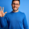 Jovem com barba vestindo suéter casual e de óculos sobre fundo azul, mostrando e apontando para cima com os dedos em número 5 enquanto sorri confiante e feliz.
