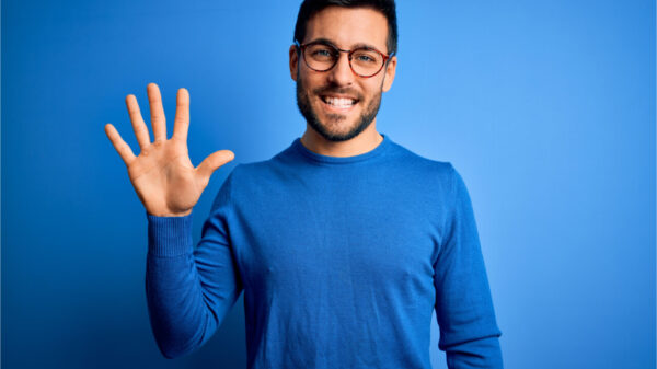 Jovem com barba vestindo suéter casual e de óculos sobre fundo azul, mostrando e apontando para cima com os dedos em número 5 enquanto sorri confiante e feliz.