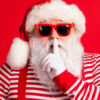 Retrato de Papai Noel misterioso de cabelos brancos, usando óculos escuros, fazendo sinal de silêncio, mudo, silencioso, isolado sobre fundo de cor vermelha.