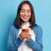 Mulher morena sorridente usando celular, isolada sobre fundo azul. Comunicação online.