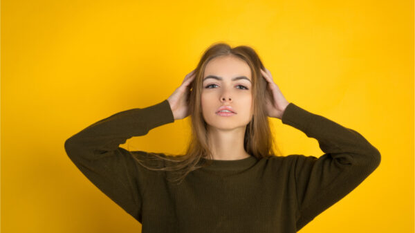 Mulher de suéter posando com os braços na cabeça e olhando para a câmera sobre fundo amarelo; foto de estúdio.