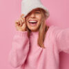 Mulher alegre e otimista usando chapeu branco e casaco, fazendo pose de expressão positiva, fazendo selfie, esticando o braço para a frente da câmera, isolada sobre fundo rosa, tirando foto de si mesma.