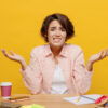 Mulher de negócios jovem, funcionária, usando camisa casual, sentada na mesa do escritório com o laptop, encolhendo os ombros, isolada no fundo de cor amarela lisa.