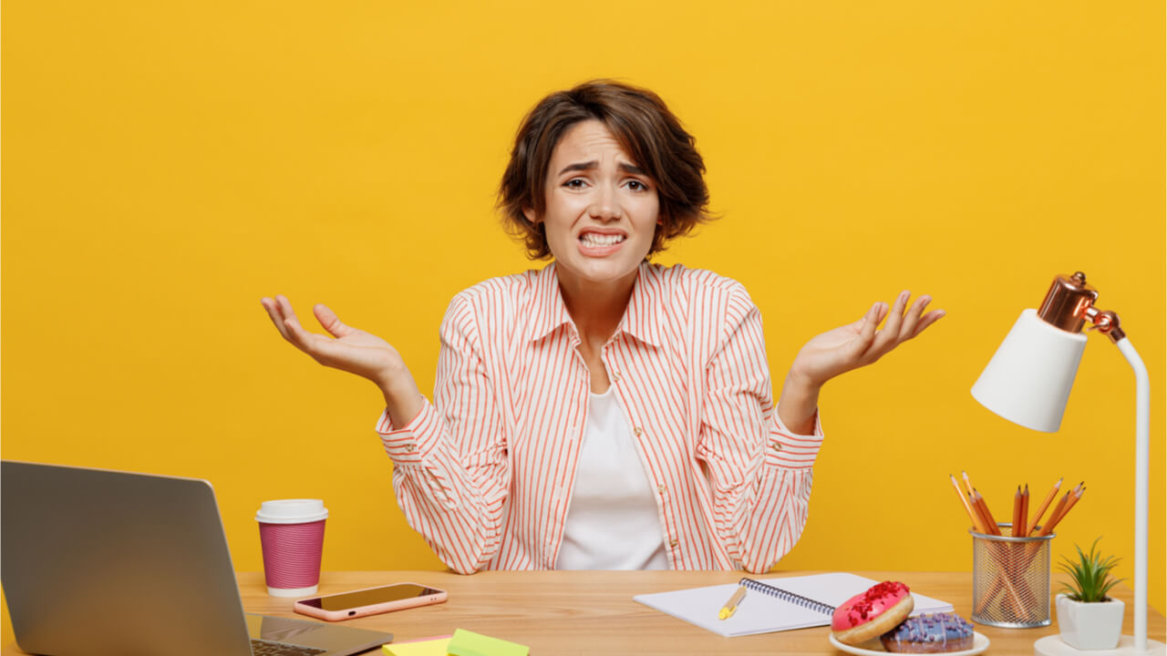 Mulher de negócios jovem, funcionária, usando camisa casual, sentada na mesa do escritório com o laptop, encolhendo os ombros, isolada no fundo de cor amarela lisa.