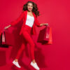 Foto de corpo todo de mulher morena saltando, fazendo compras, usando sapatos, de blazer, isolada em fundo de cor vermelha.