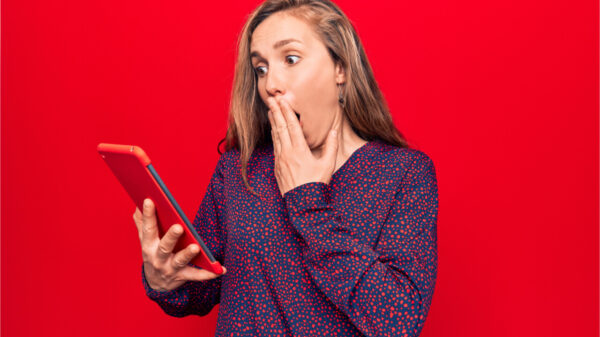 Mulher jovem usando tablet sobre fundo vermelho isolado, cobrindo a boca com a mão, chocada. Expressão surpresa.