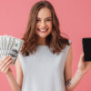 mulher segurando notas de dinheiro e um celular em fundo rosa