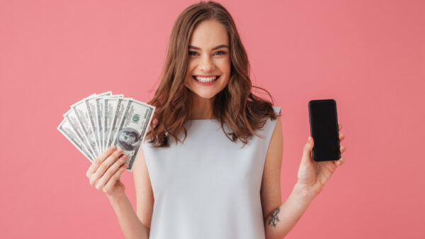mulher segurando notas de dinheiro e um celular em fundo rosa