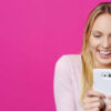 Mulher loira feliz olhando para seu celular com um sorriso surpreso contra um fundo rosa no estúdio.