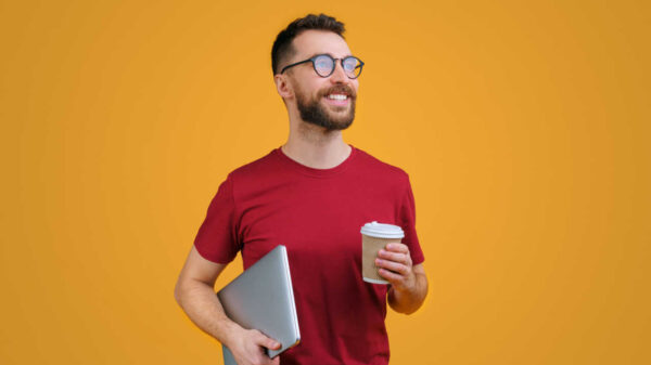 Retrato de um jovem sorridente barbudo confiante em camisetas vermelhas casuais e óculos da moda segurando laptop e xícara de café de papel, isolado em fundo amarelo.
