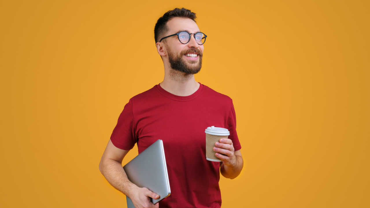 Retrato de um jovem sorridente barbudo confiante em camisetas vermelhas casuais e óculos da moda segurando laptop e xícara de café de papel, isolado em fundo amarelo.
