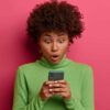 Conceito de tecnologia e emoções. Mulher estupefata usando celular moderno, olhando fixamente para tela de exibição, surpreendida, usando gola polo verde, isolada em fundo rosa.