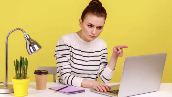 Mulher séria no escritório sentada no local de trabalho, repreendendo algo ou algém, olhando para a tela do laptop. Foto de estúdio interno isolado em fundo amarelo.