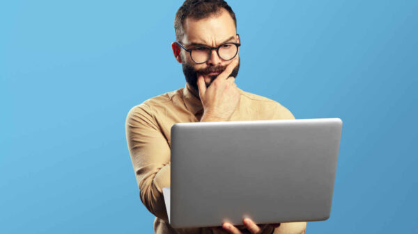 Retrato de um homem barbudo e duvidoso segurando laptop, isolado sobre fundo azul.