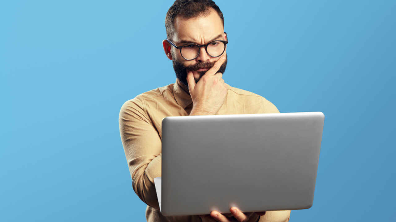 Retrato de um homem barbudo e duvidoso segurando laptop, isolado sobre fundo azul.