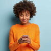 Foto horizontal de uma adolescente satisfeita, focada em dispositivo de smartphone, usando suéter laranja casual.