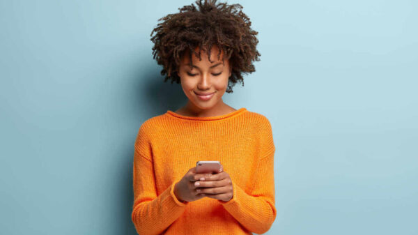 Foto horizontal de uma adolescente satisfeita, focada em dispositivo de smartphone, usando suéter laranja casual.