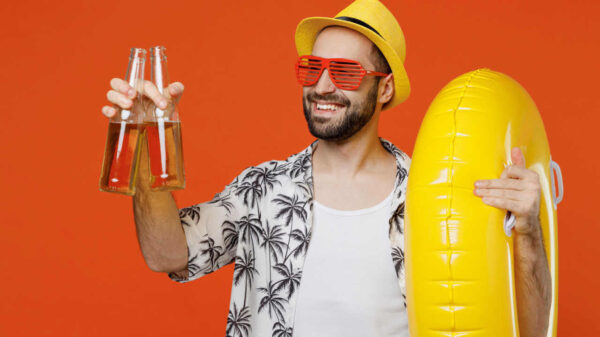 Jovem sorridente usando chapéu, camisa de praia e óculos, segurando garrafas de cerveja e boia inflável, olhando para o lado, isolado no retrato de estúdio de fundo laranja simples.