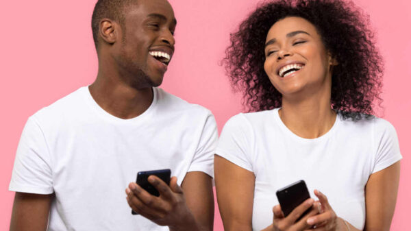 Casal jovem e feliz rindo e usando smartphones isolados no fundo do estúdio rosa. Amigos alegres segurando aparelhos de telefones celulares e se divertindo com celulares juntos.