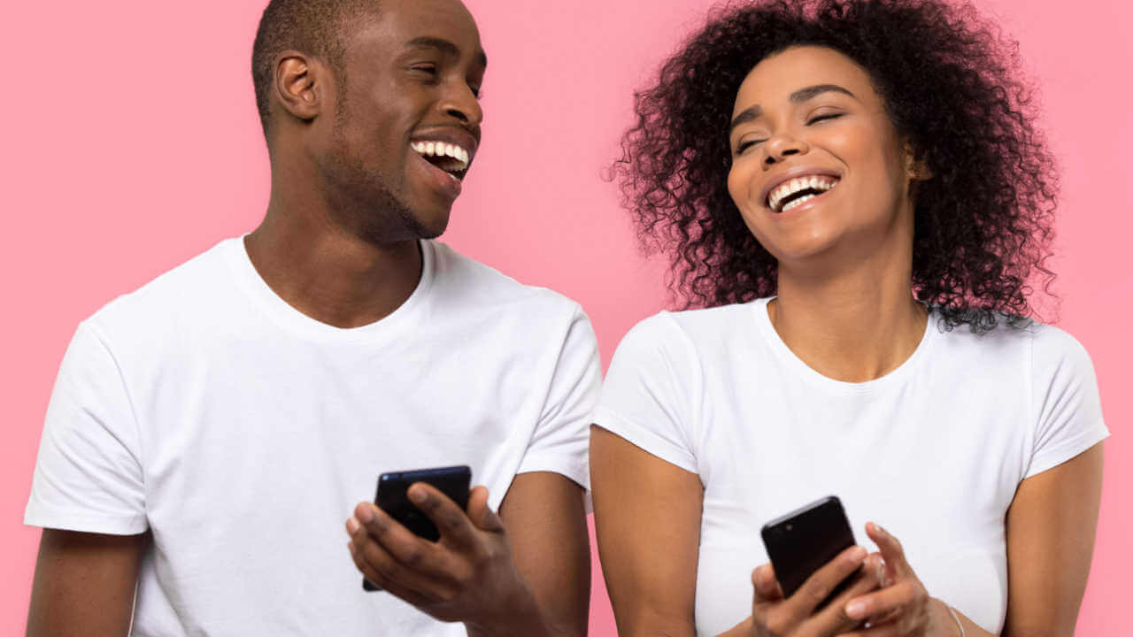 Casal jovem e feliz rindo e usando smartphones isolados no fundo do estúdio rosa. Amigos alegres segurando aparelhos de telefones celulares e se divertindo com celulares juntos.
