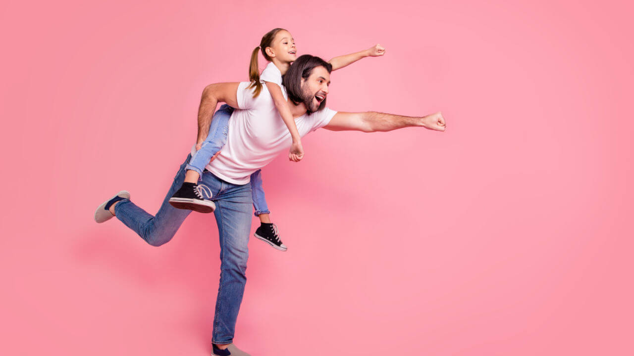 Foto de corpo de menina com o pai a segurando nas costas, e os dois com o braço esticado, usando camisetas brancas casuais e calças jeans, isolados no fundo rosa.