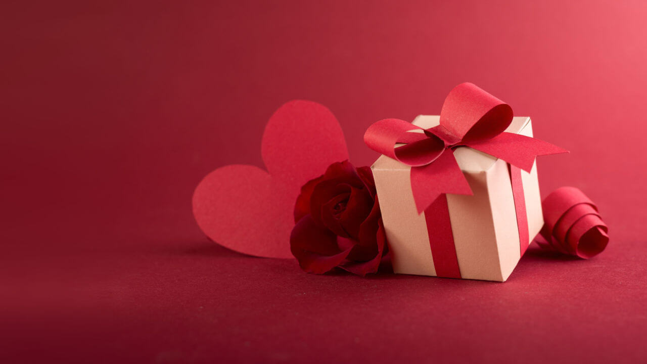 Arte em papel com caixa de presente feita à mão, fita de corte de papel, flor e coração em um fundo vermelho.