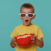 Menino feliz de camiseta comendo pipoca e assistindo a filme, usando óculos 3D, desfrutando de pipoca. Criança isolada sozinha no fundo azul do estúdio.