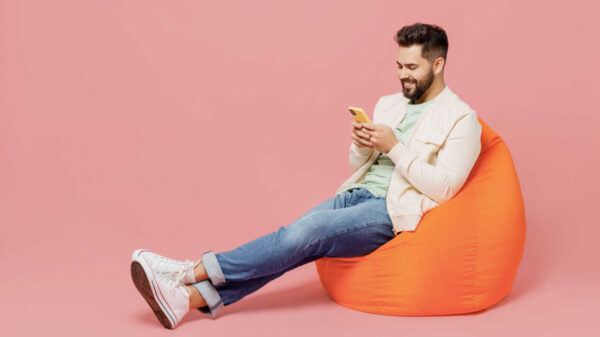homem sentando num puff mexendo no celular em fundo rosa - Foto: Shutterstock